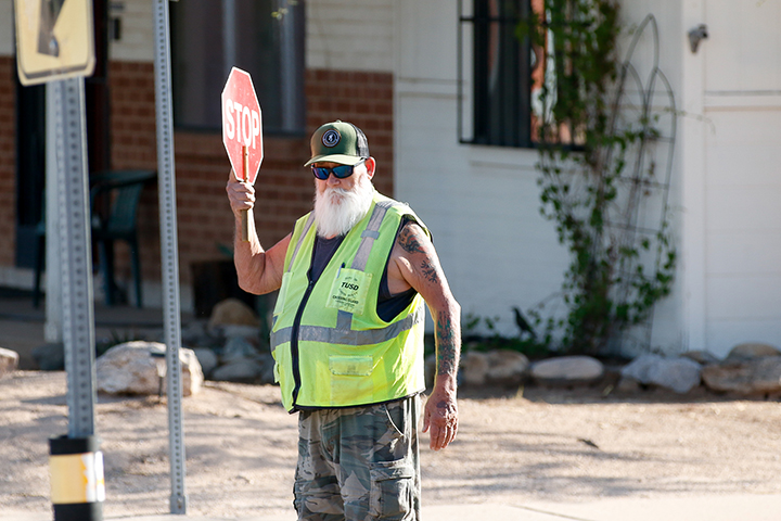 A man with a white beard wearing a yellow safety vest and holding a stop sign walks across the street